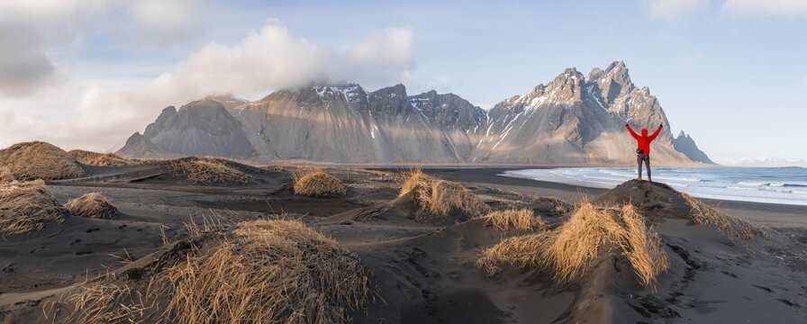Man in red jacket stretches arms into the air, black lava beach, sandy beach, dunes with dry grass, mountains Klifatindur, Eystrahorn and Kambhorn, headland Stokksnes, mountain range Klifatindur, Austurland, East Iceland, Iceland