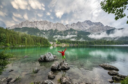 Young man sticks his arms in the air and stands on a rock on the shore, Eibsee lake in front of Zugspitze massif with Zugspitze, dramatic Mammaten clouds, Wetterstein range, near Grainau, Upper Bavaria, Bavaria, Germany