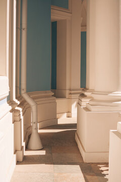 Interior view of a classical colonnade featuring tall white columns, light blue walls and elegant architectural details. A white drainpipe runs along the wall while strong natural sunlight casts long 
