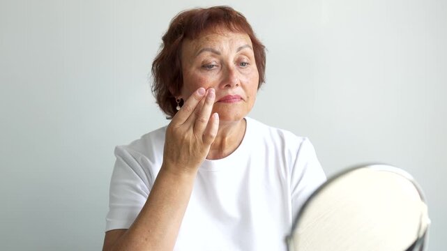 Adult woman examines wrinkles in a small mirror indoors. An older woman looks at reflection in front of mirror and touches her face with her hands. Concepts of beauty, care, and aging.