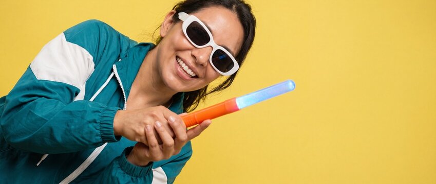 Smiling woman holding glowing stick in teal retro windbreaker jacket and white sunglasses posing against bright yellow background wall playing with lightup toy wand enjoying party rave neon energy