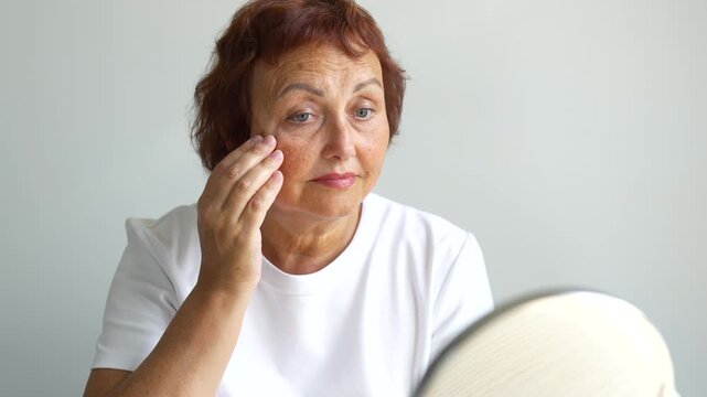Adult woman examines wrinkles in a small mirror indoors. An older woman looks at reflection in front of mirror and touches her face with her hands. Concepts of beauty, care, and aging.