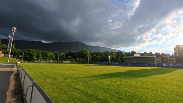 Sofia, Bulgaria - 06.06.2026. Wide panoramic view of DIT Dragalevtsi Stadium with natural green grass football field and grandstands under dramatic storm clouds. Mountains visible in the background