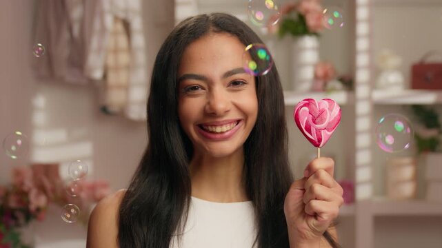 Portrait happy smiling American girl holding candy love heart shape in pink bathroom flying soap bubbles close up positive Hispanic young woman Latina ethnic female smile hold sweet sugar diet problem