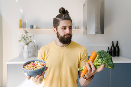 Confused man holding vegetable and pills bowl at home
