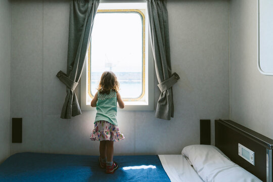 Back view of little girl standing on bed looking to the sea from window of ship's cabin