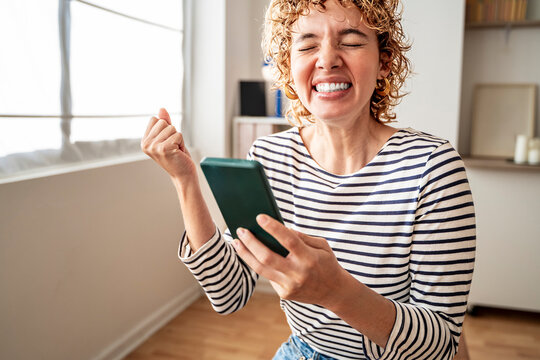 Excited woman celebrating winning while using smartphone indoors