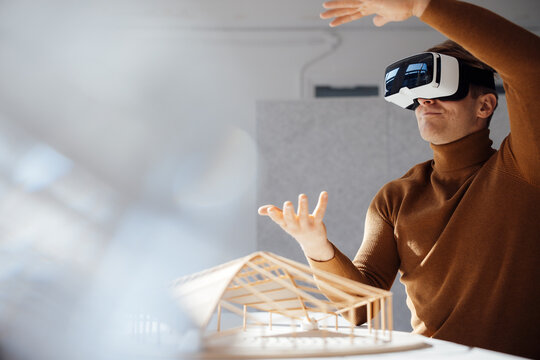 Architect wearing virtual reality simulator gesturing sitting with leaf shaped model at desk in office