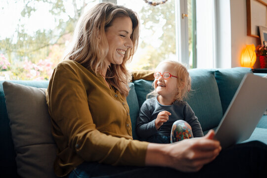 Cheerful blond woman holding tablet PC sitting by daughter on sofa at home