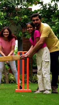 Indian family playing cricket batting in home garden, spending quality fun time together playing outdoor sports, cheerful Asian parents and children enjoying backyard activity bonding on sunny day
