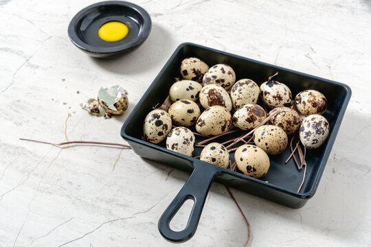 Fresh quail eggs in square frying pan and cracked quail eggshell with yolk, on marble table