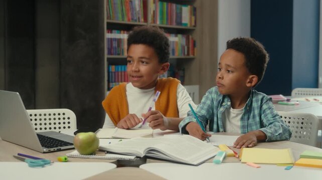 E-learning distant primary education two African American little boys watching lesson with laptop technology web study writing together children kids schoolboys pupils sons write studying at school
