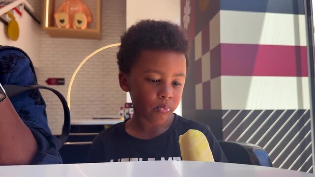 close-up view of a 6-yer-old biracial black child wearing curly hair and expressive black eyes eating a vanilla ice cream sitting next to his mother inddor