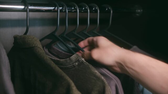 Male hand selecting t shirt from variety colorful clothes hanging on hangers in dark wardrobe, carefully making choice for day outfit before pulling one out. Man choosing clothes from dark closet