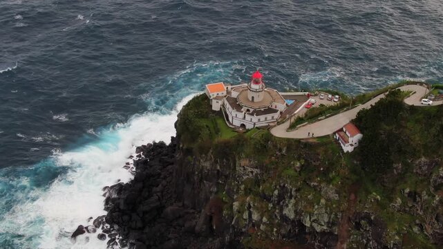 Aerial perspective of the Ponta do Arnel lighthouse standing on a steep cliff while powerful ocean waves crash against the rocky coast