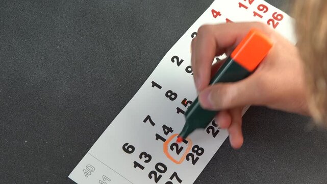 Hand Marking an Important Date on a Calendar Page with an Orange Highlighter. Planning date. Circle and strike through calendar. Write