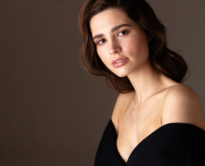 Naklejka premium Beauty portrait of a young woman with wavy brunette hair wearing an elegant black off-shoulder dress. Natural makeup, clear skin, and a confident gaze create a sophisticated fashion concept.