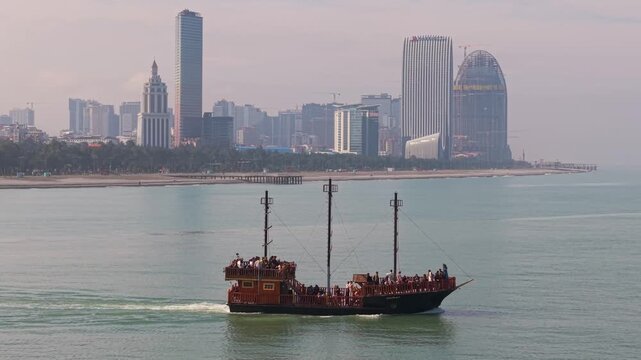 Drone footage follows a wooden pirate-themed tourist boat cruising the Black Sea, framed against the striking modern skyscrapers of the Batumi city skyline.