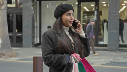 Fototapeta premium Young latin woman in beret holds colorful shopping bags and phone to ear while standing outside a retail store on a busy street; leisure contentment.