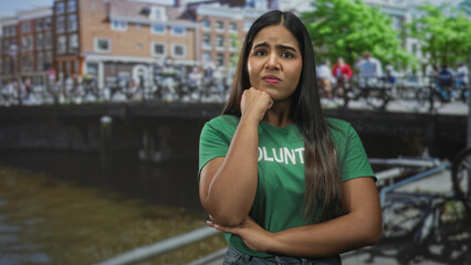 Fototapeta premium Young woman volunteer with hand on chin and crossed arms on a street by a canal and bicycles; concern reflection.