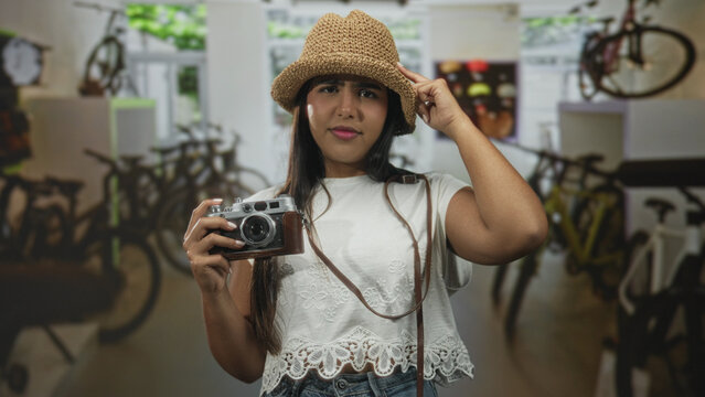 Young hispanic woman holding film camera and points finger toward viewer in a bike shop inside a building; travel inspiration playful.