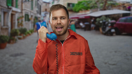 Fototapeta premium Man prisoner in orange jumpsuit holding blue handset to ear on a street with cobblestones and blurred cafe stalls visible; isolation regret.