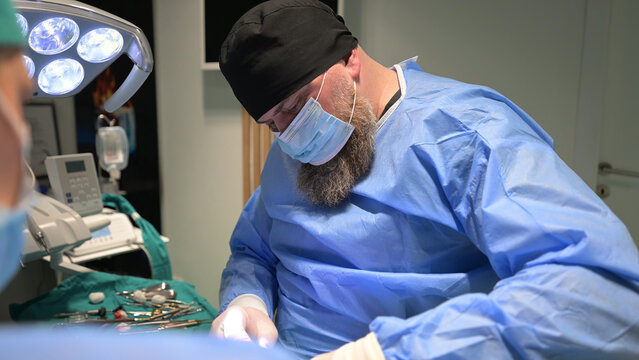 Bearded Surgeon Performing Operation in Modern Operating Room Wearing Surgical Mask and Gown
