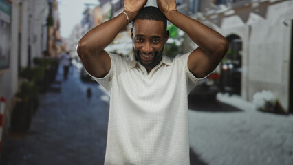 Naklejka premium Man with hands above head and palms raised on street wearing white polo shirt, smiling toward camera; playful moment.