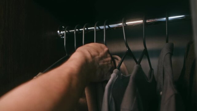 Close up person hand selecting outfit from dark wardrobe, carefully browsing through casual clothing hanging on rail before making choice for day ahead. Person choosing clothes from a dark home closet