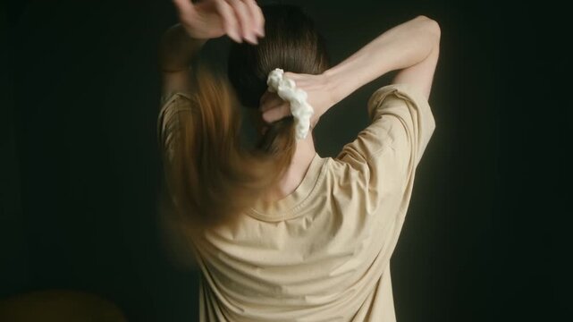 Rear view brunette woman gathering long healthy brown hair, tying itin low ponytail with white scrunchie, dark background, showcasing simple hairstyle routine. Woman with long hair making a ponytail