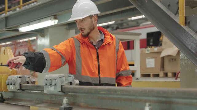 Orange-vested technician meticulously examines electric train machinery within dimly lit factory setting featuring conveyor belts and industrial lighting fixtures for maintenance inspection.