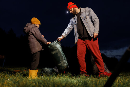 Young bearded father with headlamp flashlight packing sleeping bag or camping gear with little son on lake shore at dark night. Caring parent and boy setting up camp outdoors during autumn family acti