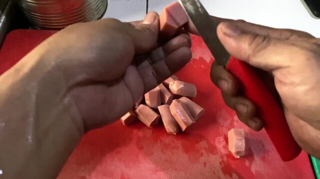 Hands slicing raw sausages on a cutting board A descriptive close-up stock footage clip showing a person's hands using a sharp kitchen knife to slice raw sausages into pieces on a cutting board. 