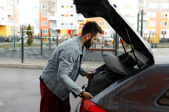 Young bearded man loading black luggage bag into car trunk on city street. Casual male driver packing suitcase in open hatchback automobile boot outdoors. Everyday commuter lifestyle before road trip