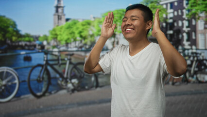 Fototapeta premium Man raising hands and smiling on street near an amsterdam canal with bicycles and water, wearing white tshirt and closed eyes; joy.