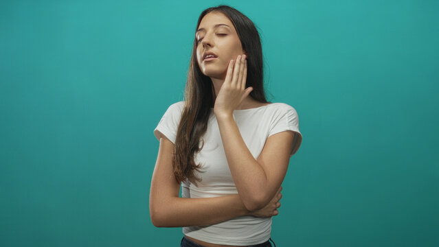 Young hispanic woman with eyes closed, hand on neck and left arm across waist in teal studio; serenity.