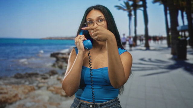 Woman wearing blue holding vintage telephone handset points finger on street promenade by ocean and palm trees; impatience.