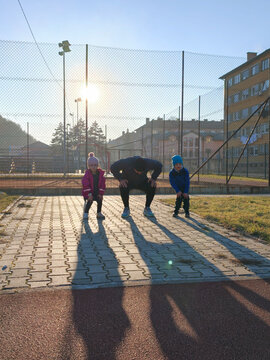 Father Coaching Children Doing Warm-Up Squats On Outdoor Sports Court At Sunset