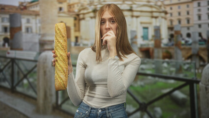 Fototapeta premium Woman holding a baguette with fingers to mouth gesture at building piazza near ancient railing and fountain; pensive hunger hesitation.