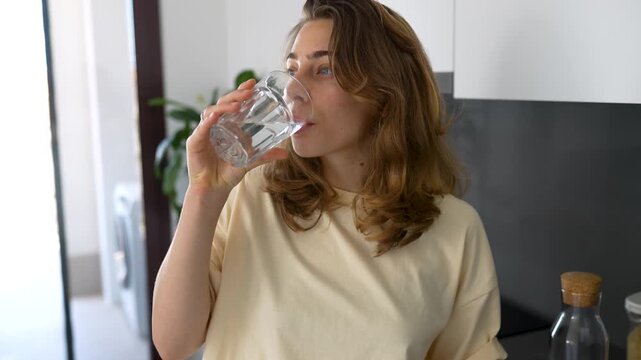 A woman drinks water from a clear glass in a modern kitchen. A young woman enjoys a sunny day, pouring water from a carafe in a sunny room. Concepts of beverages, enjoyment, and homeliness.