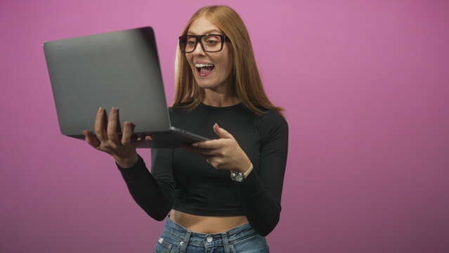 Woman holding laptop, smiling and waving hand with midriff visible in studio pink backdrop, wearing glasses and jeans; virtual meeting joy.