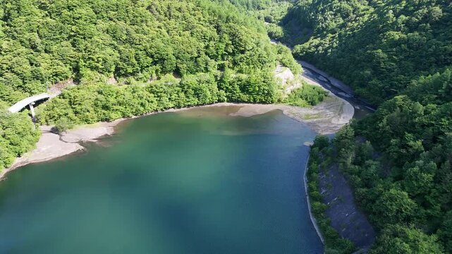 Aerial view of emerald river winding through lush green hills. Concrete bridge spans the flowing water's curve. Riverside banks show sandy patches amid dense foliage