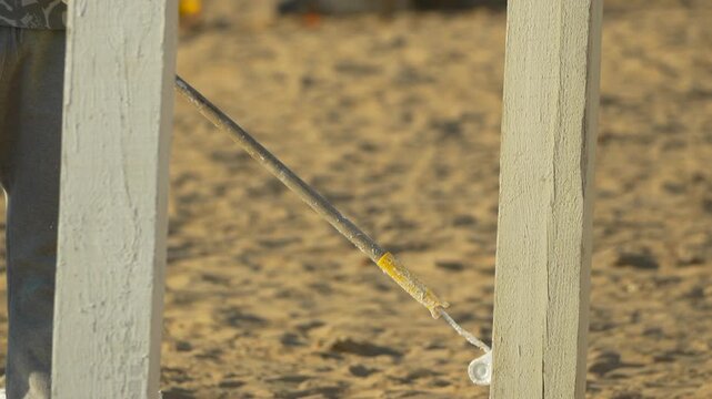 Painting wood beach worker uses a long handled roller to apply white paint to a wooden post on a sandy shore