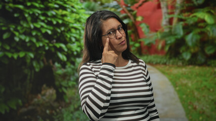 Naklejka premium Woman pointing finger and pursing lips wearing glasses beside a building walkway with green shrubs; skepticism reflection thought.