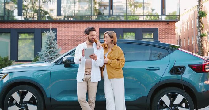 Young couple using tablet while electric car charges in parking lot