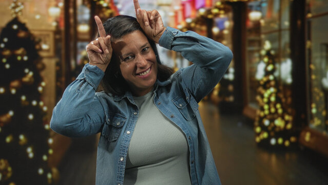 Woman making antlers with fingers and smiling amid holiday lights and decorated storefronts inside a shopping mall building; playful holiday joy.