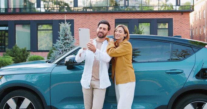 Young couple video calling friends while charging electric car on road trip