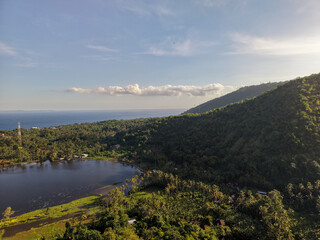 Naklejka premium Aerial panoramic landscape of a coastal lake and forested mountain slopes facing the ocean in Karangasem, Bali