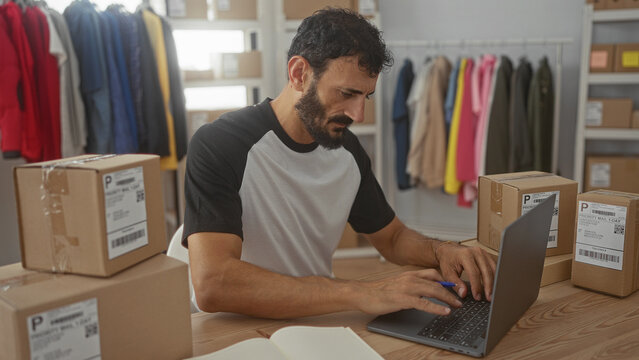 Man writing with pen by laptop and stacked shipping boxes, hand on keyboard while labeling parcels inside building; focus entrepreneurship.