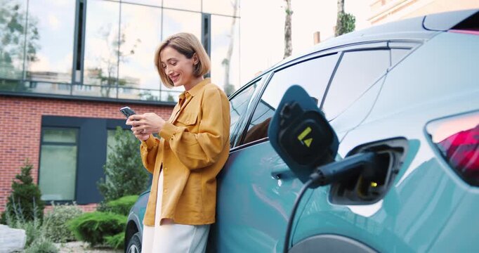 Adult woman using mobile app to charge an electric car at a parking lot outdoors.
